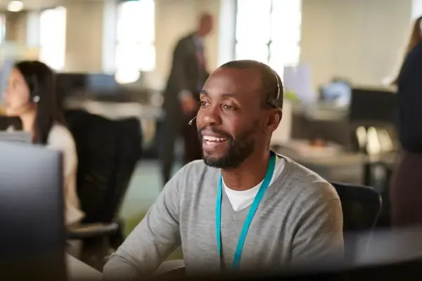 A smiling male call center employee wearing a headset looks at his computer screen in a busy open-plan office.